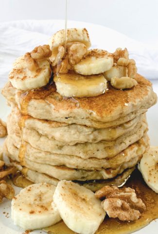 Stack of pancakes topped with cut up banana pieces, walnut pieces, and syrup drizzling onto them
