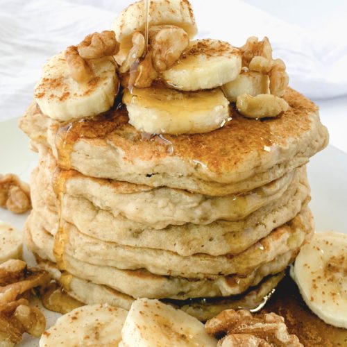 Stack of pancakes topped with cut up banana pieces, walnut pieces, and syrup drizzling onto them