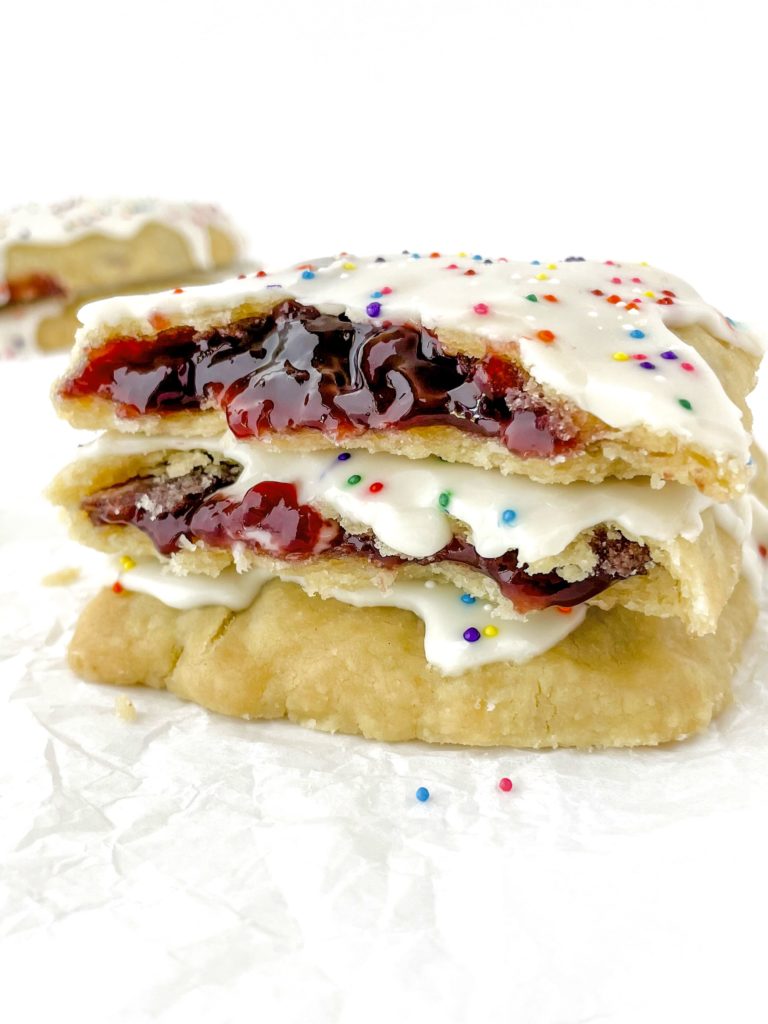 Photo of a stack of 3 homemade vegan pop tarts each with white icing and rainbow sprinkles, and two of them broken open with red jam insides