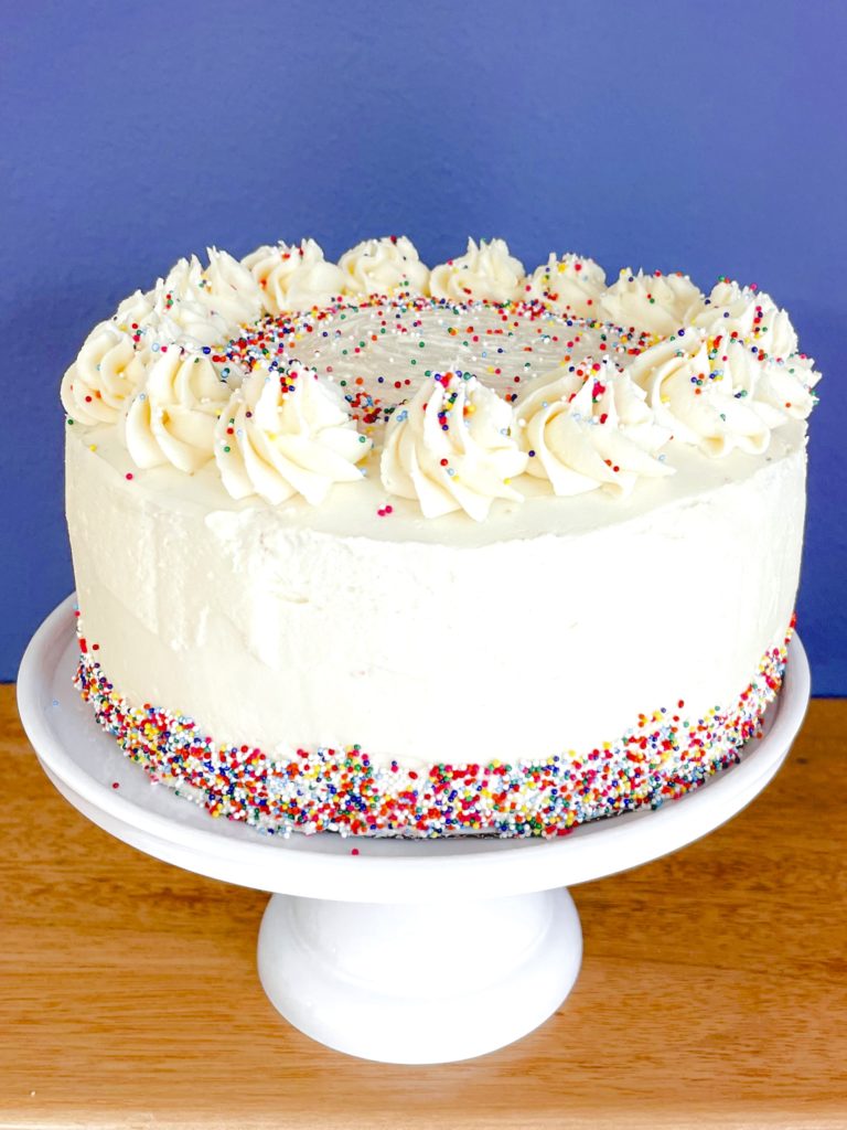 Vegan Funfetti Cake - looking down at the full cake on a white cake stand with a purple backdrop and wooden counter