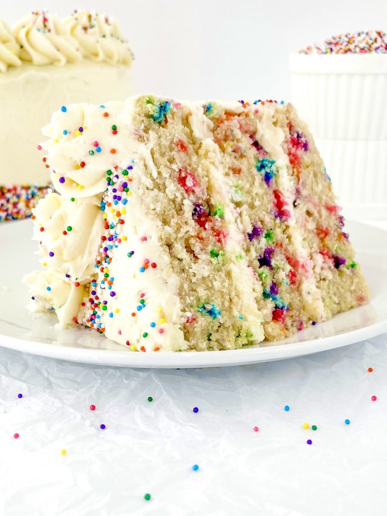 Vegan Funfetti Cake - one huge chunky slice on a white plate and the full cake and more sprinkles in the background
