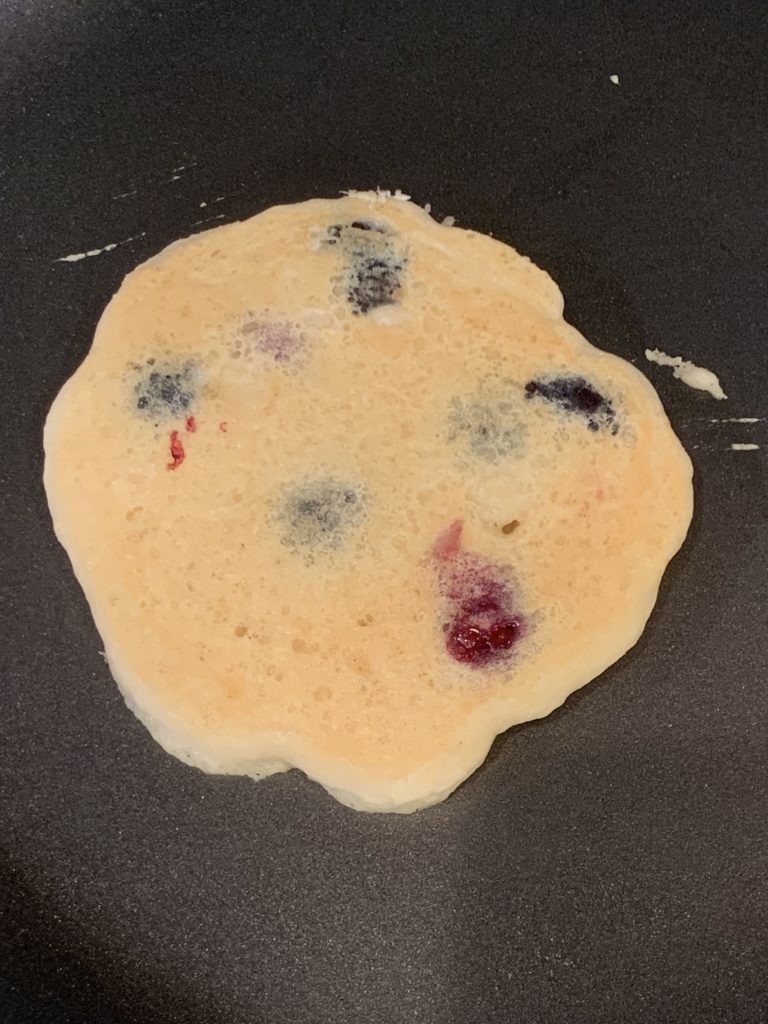 One blueberry pancake with the top half cooked, on the pan