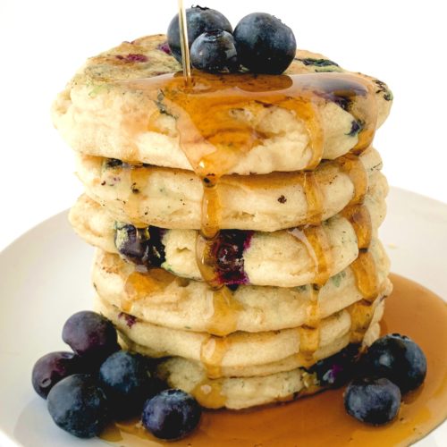 Big stack of super fluffy blueberry pancakes topped with fresh berries with maple syrup drizzling down onto them