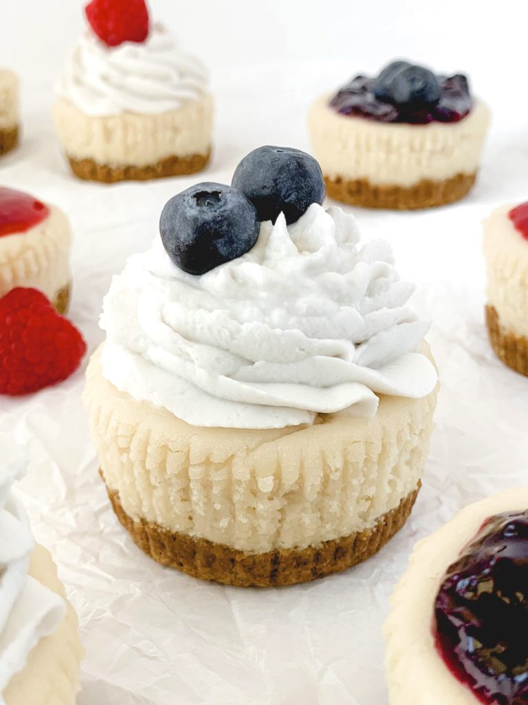 Lots of mini cheesecakes on a white background, the closest one topped wit whipped cream and fresh blueberries