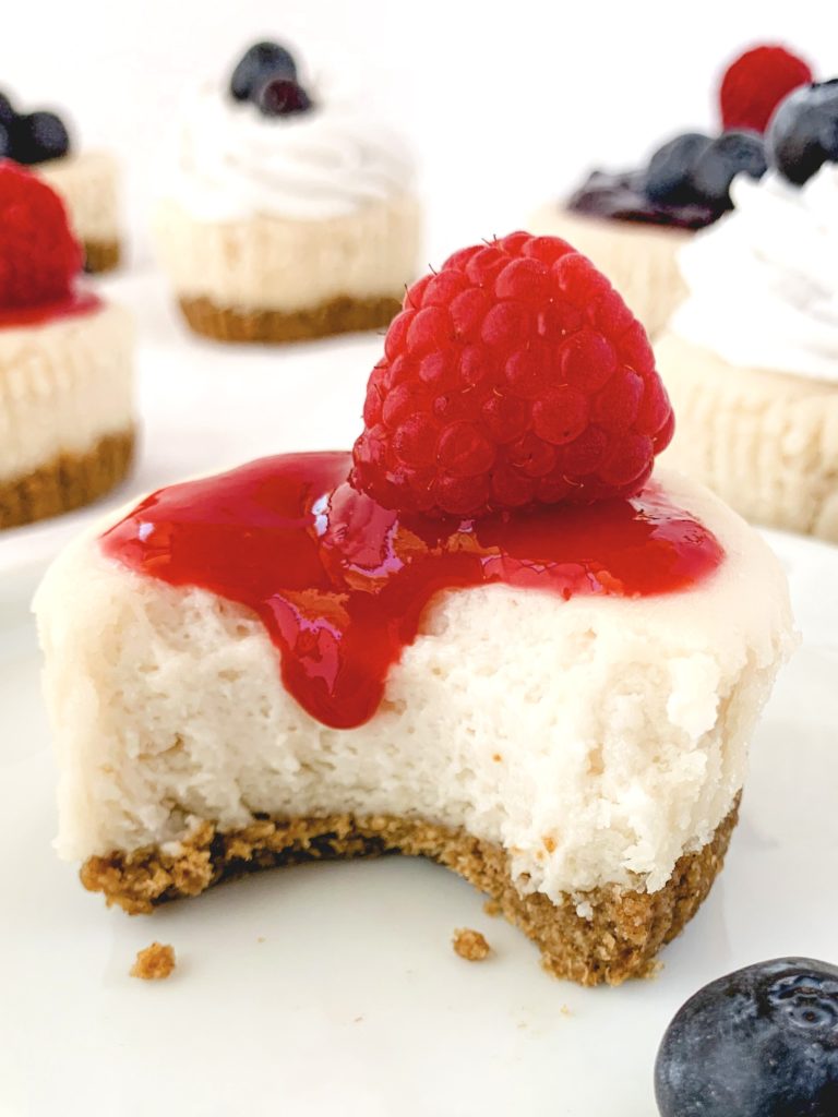 Close up of one mini cheesecake with a bite out of it, topped with raspberry sauce and one fresh raspberry
