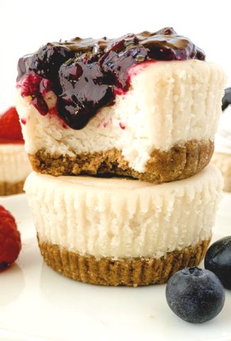 Two mini cheesecakes stacked on one another, the top one with a bite taken out, and topped with a dripping blueberry compote