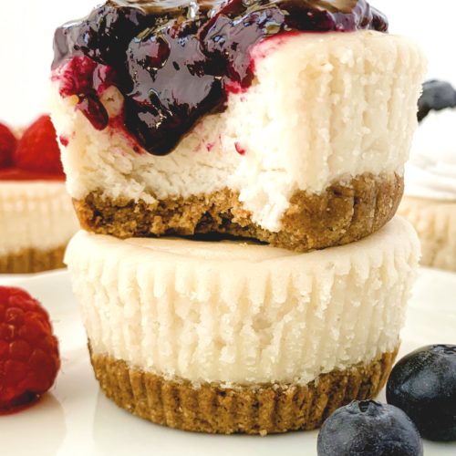 Two mini cheesecakes stacked on one another, the top one with a bite taken out, and topped with a dripping blueberry compote